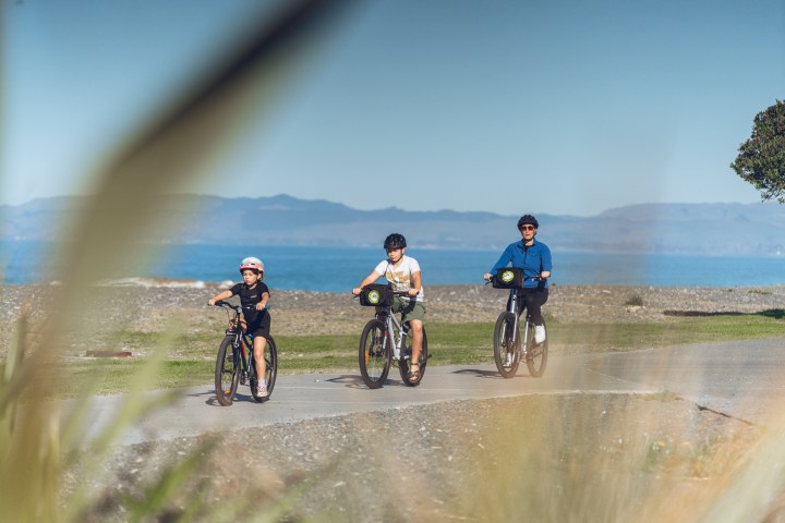 Three people biking on a path by the sea with distant mountains.