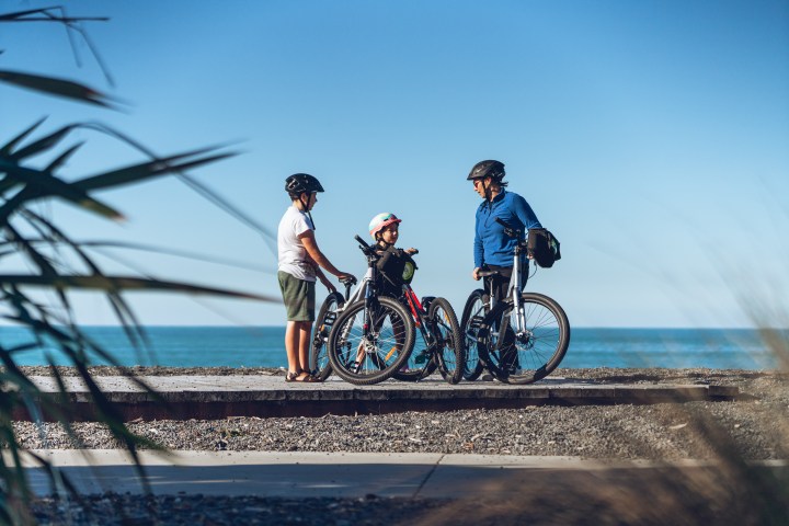 Three people with bikes near a railway track by the sea under clear blue sky.