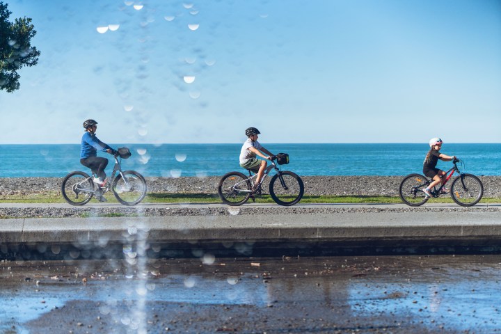 Three kids biking by the sea on a sunny day, water droplets in the foreground.