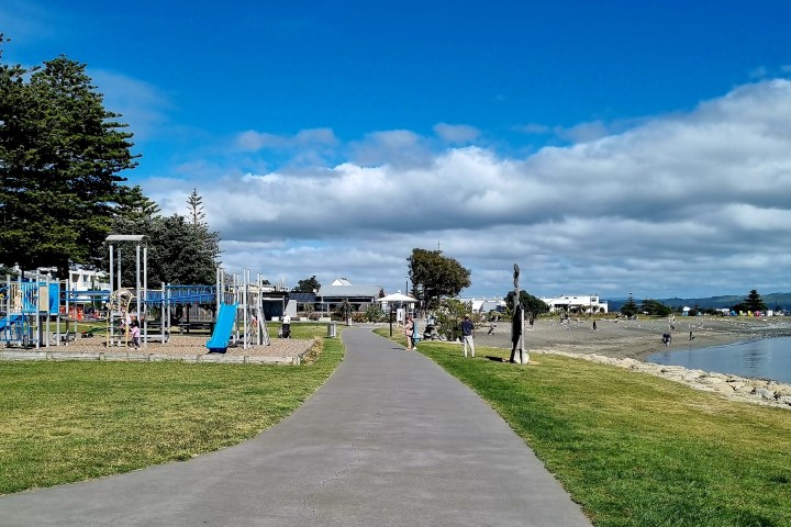 Pathway along beach with playground, trees, and people under a blue sky.