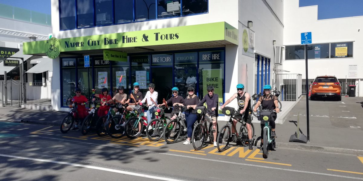 Group of cyclists outside Napier City Bike Hire & Tours building on a sunny day.