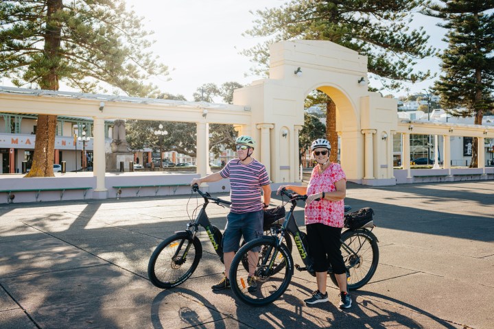 Two people with bikes stand in front of an arch in a sunny plaza with trees.