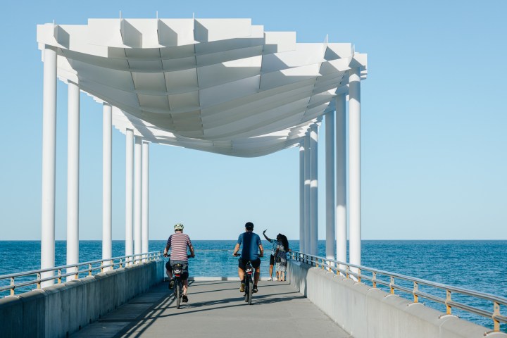 Two cyclists and a pedestrian on a pier with white canopy over the ocean.