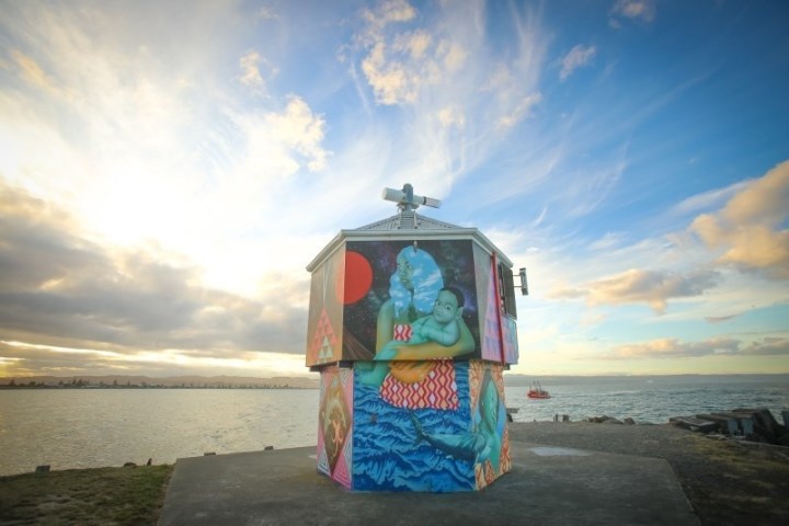 Art-covered structure by water, featuring colorful mural under cloudy sky.
