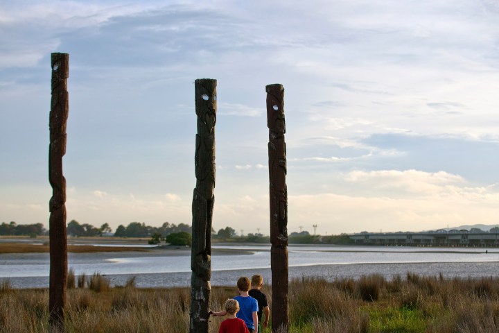 Three kids explore tall wooden poles by a lakeshore under a cloudy sky.